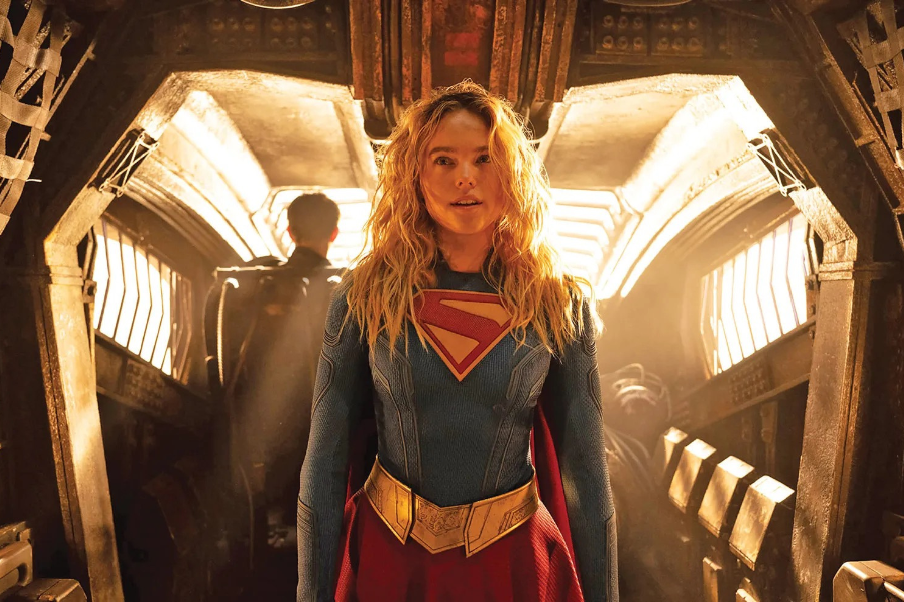Supergirl Review: Strong Performances Can’t Save an Uneven Adventure Supergirl in a blue suit with a red cape and gold belt stands in a dim, industrial spaceship corridor.