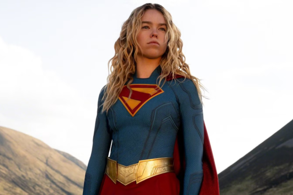 Supergirl standing outdoors in a blue suit with a red cape and gold belt, mountains in the background.