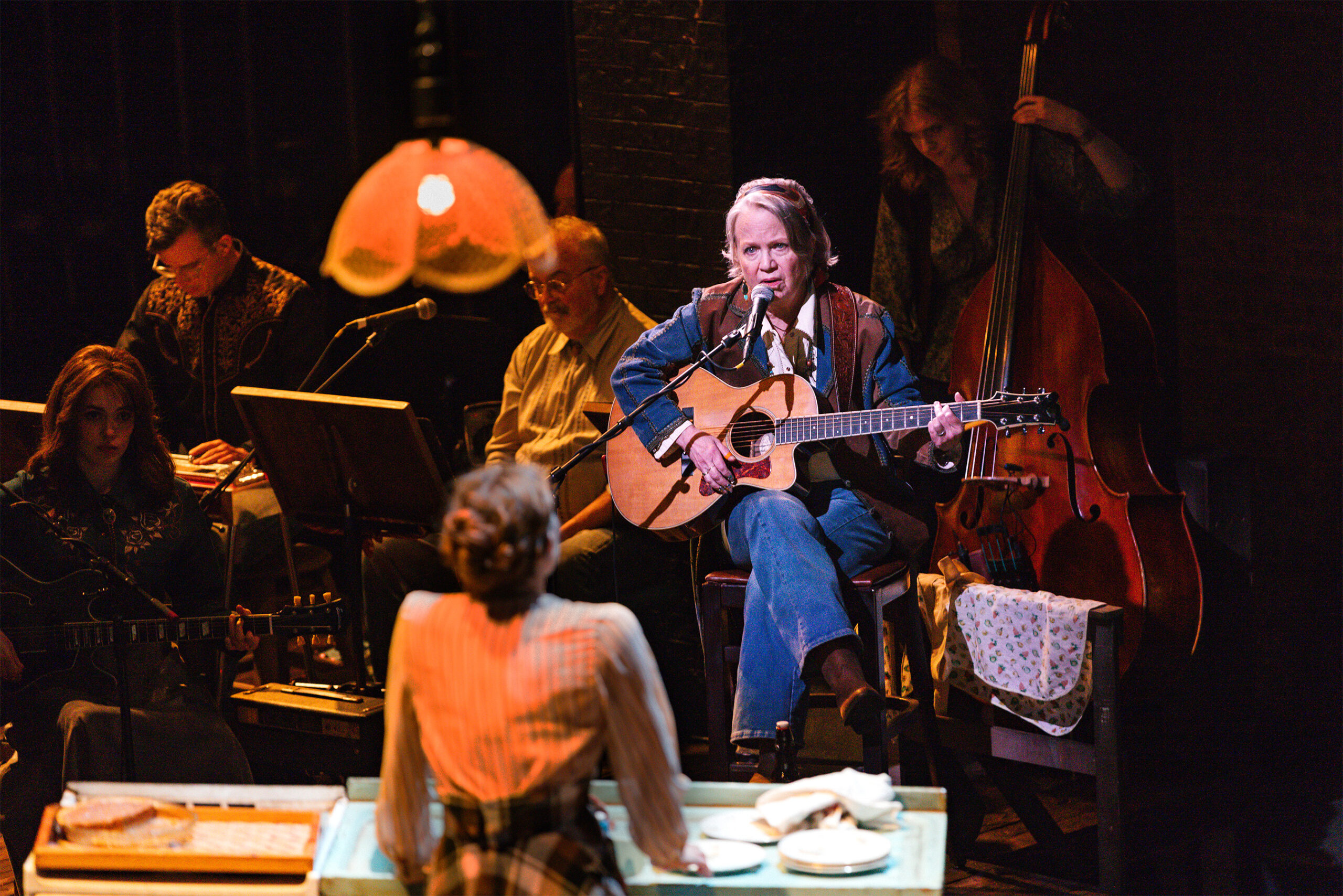 6_CST_BROKEBACK_Press_KyleFlubacker - Eulalie Magazine Chicago Shakespeare Theater’s Brokeback Mountain Review Female guitarist and vocalist center stage, singing into a microphone with a small band in a dimly lit venue.