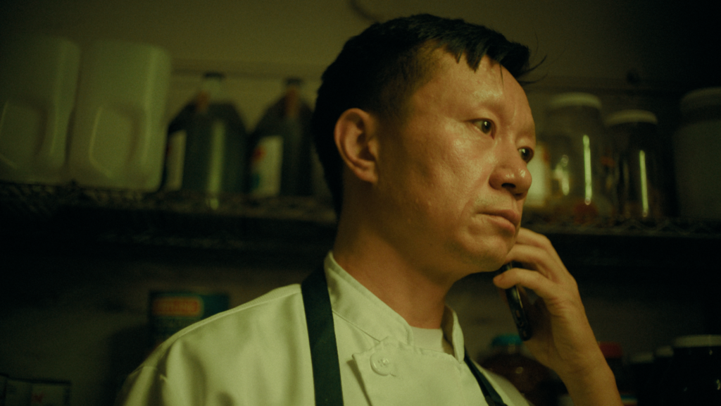 Benny Chen Discusses His Short, HAND Man wearing a white chef's coat and apron, talking on a phone in a dimly lit kitchen.