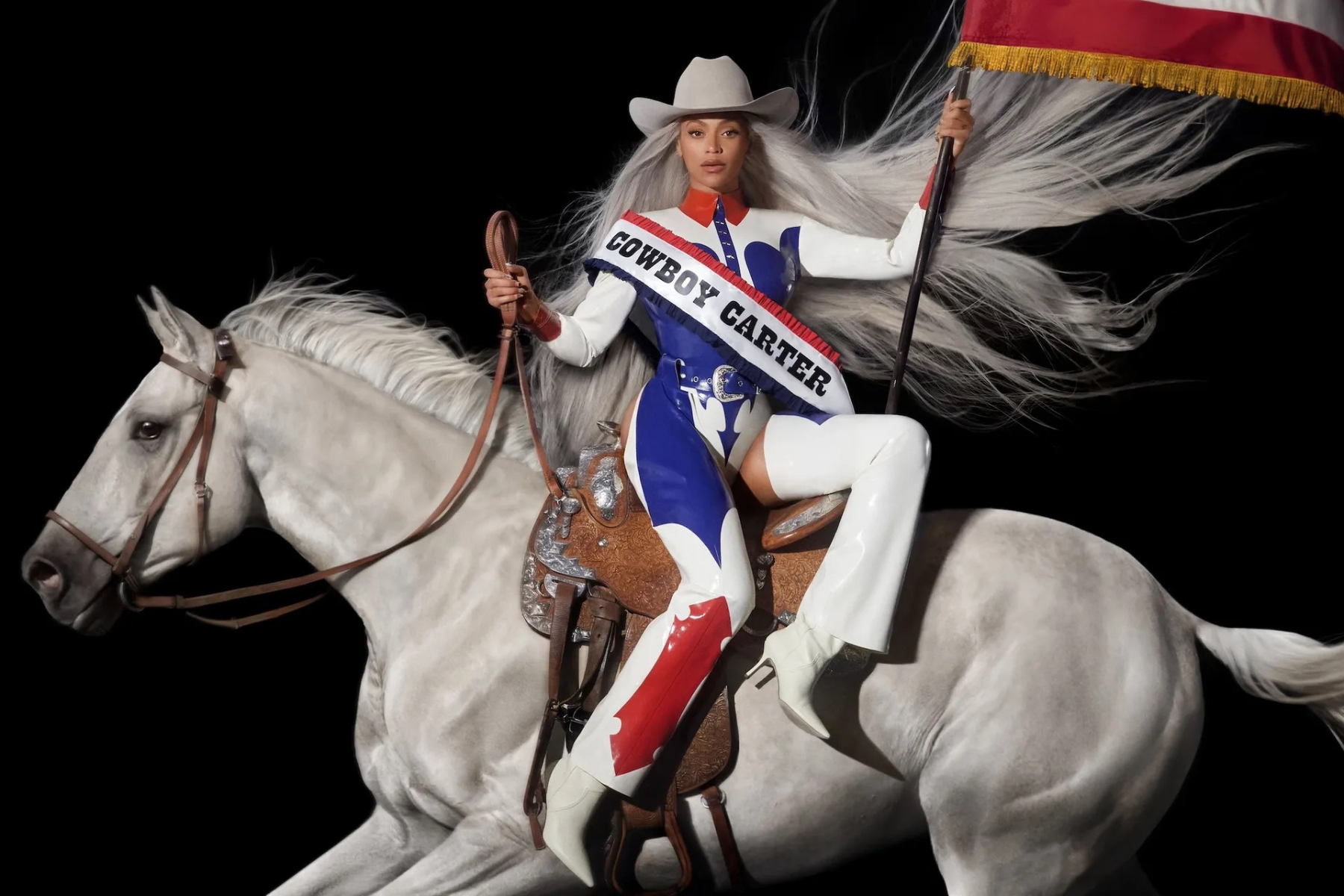 Every Beyoncé Album – Ranked! Female rider in a white hat and blue-red-silver costume atop a white horse, waving a flag against a black background.