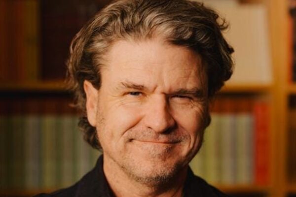 Smiling man with wavy gray hair in a dark shirt, standing in front of a bookshelf filled with books
