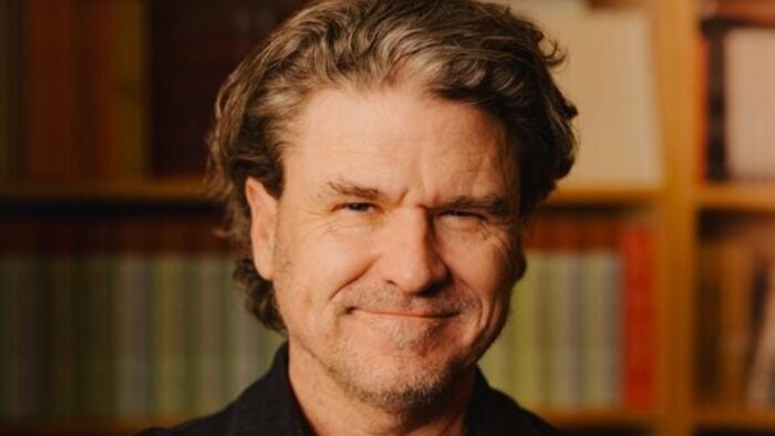 Chicago Humanities’ Dave Eggers Discussion Review Smiling man with wavy gray hair in a dark shirt, standing in front of a bookshelf filled with books