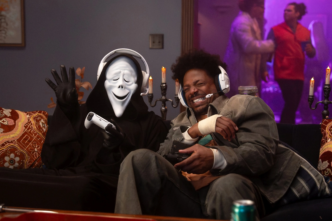 Ghostface - Eulalie Magazine Scary Movie 6 Review: Only a Few Zany Scenes Hit the Comic Bullseye in This Wild Sequel A masked figure in a black cloak wearing a headset waves while holding a game controller next to a laughing man with headphones on a living room couch, gaming at home.