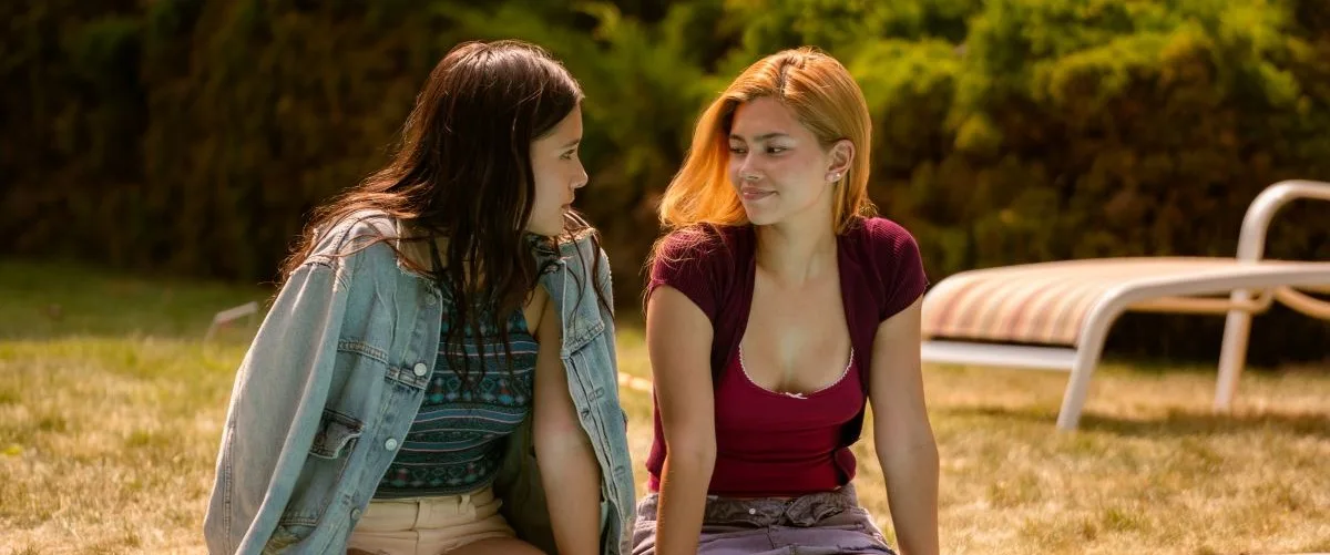 Girls Like Girls Review: Heartbreak Feels Good in a Movie Like This Two young women sit on a sunlit grassy lawn, facing and chatting with smiles on their faces in a park setting.