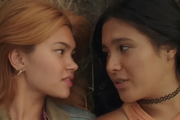 Two women face each other closely outdoors, one with light brown hair and the other with black hair, engaged in conversation.
