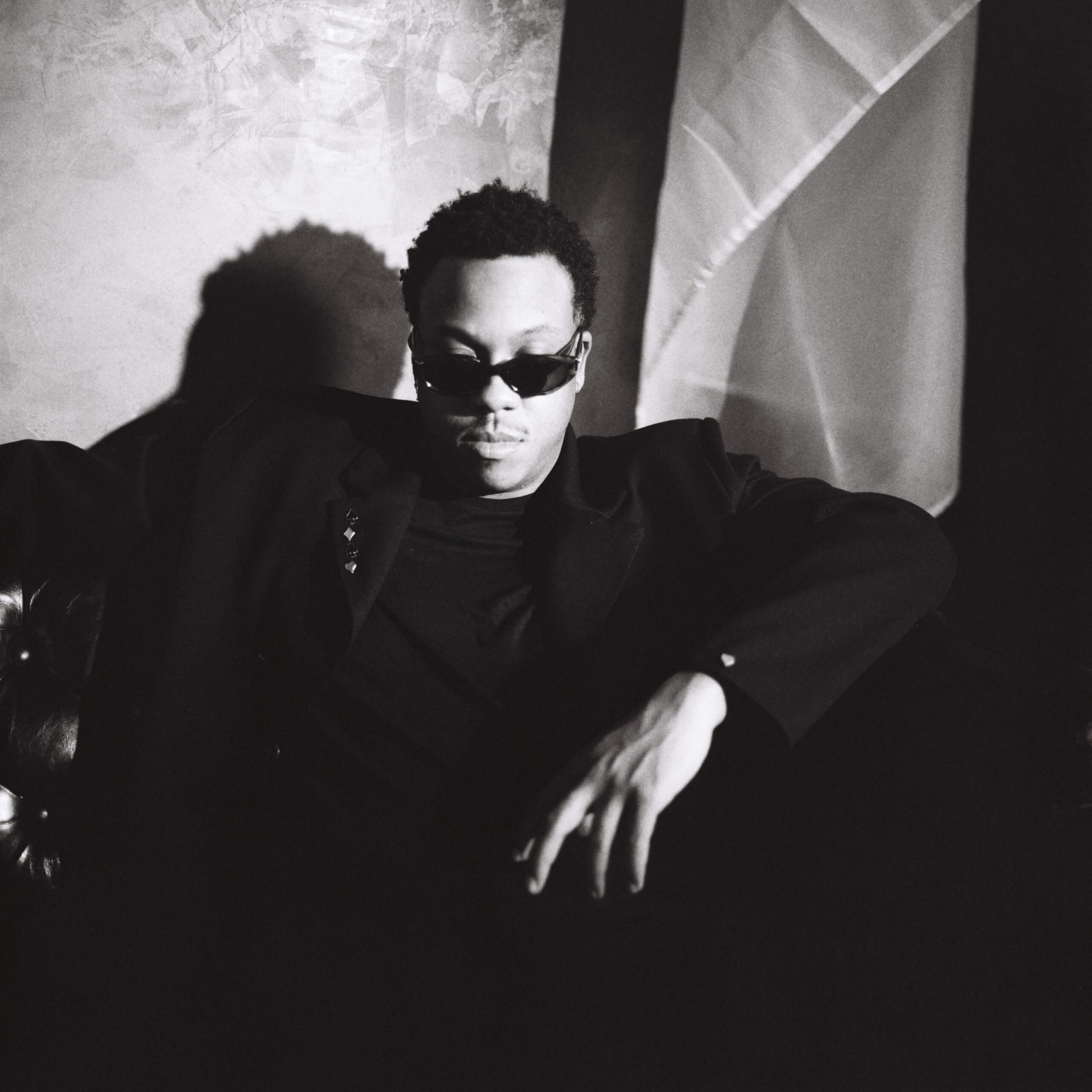 Lee Lewis on New EP, HOWL, Broken Relationships, and Healing Black-and-white portrait of a person with short curly hair wearing sunglasses, arms extended outward against a textured backdrop.