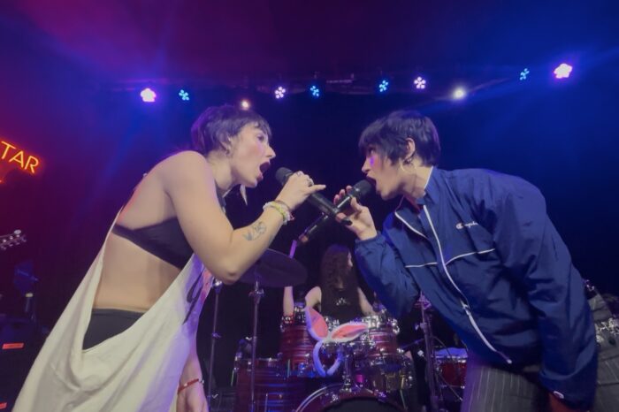 Daisy Grenade’s First Headline Tour is an Explosive, Memorable Debut Two female performers sing into handheld microphones on a lit stage, facing each other, with a drum kit and a neon Mortar Hall sign in the background.