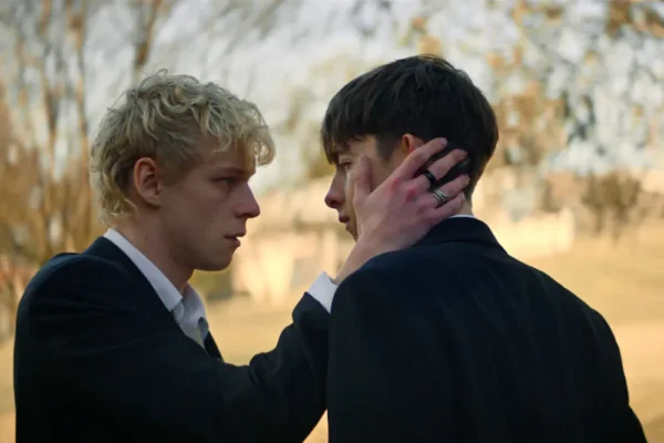 Two men in dark suits face each other; one cups the other's cheek in a tender moment outdoors.