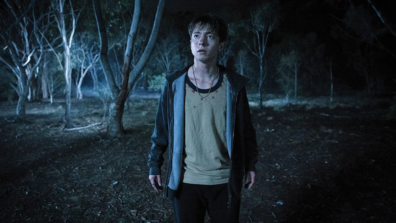 Leviticus Review: A Frightening Horror Movie That Has a Lot on Its Mind Young man stands in a dark forest at night, wearing a beige shirt and hoodie, looking frightened.
