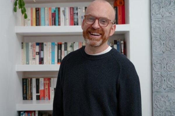 Smiling man with glasses and a red beard standing in front of white bookshelves in a home library setting.