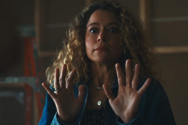 Woman with curly blonde hair raises both hands, eyes wide, in a dimly lit room, showing surprise or fear.
