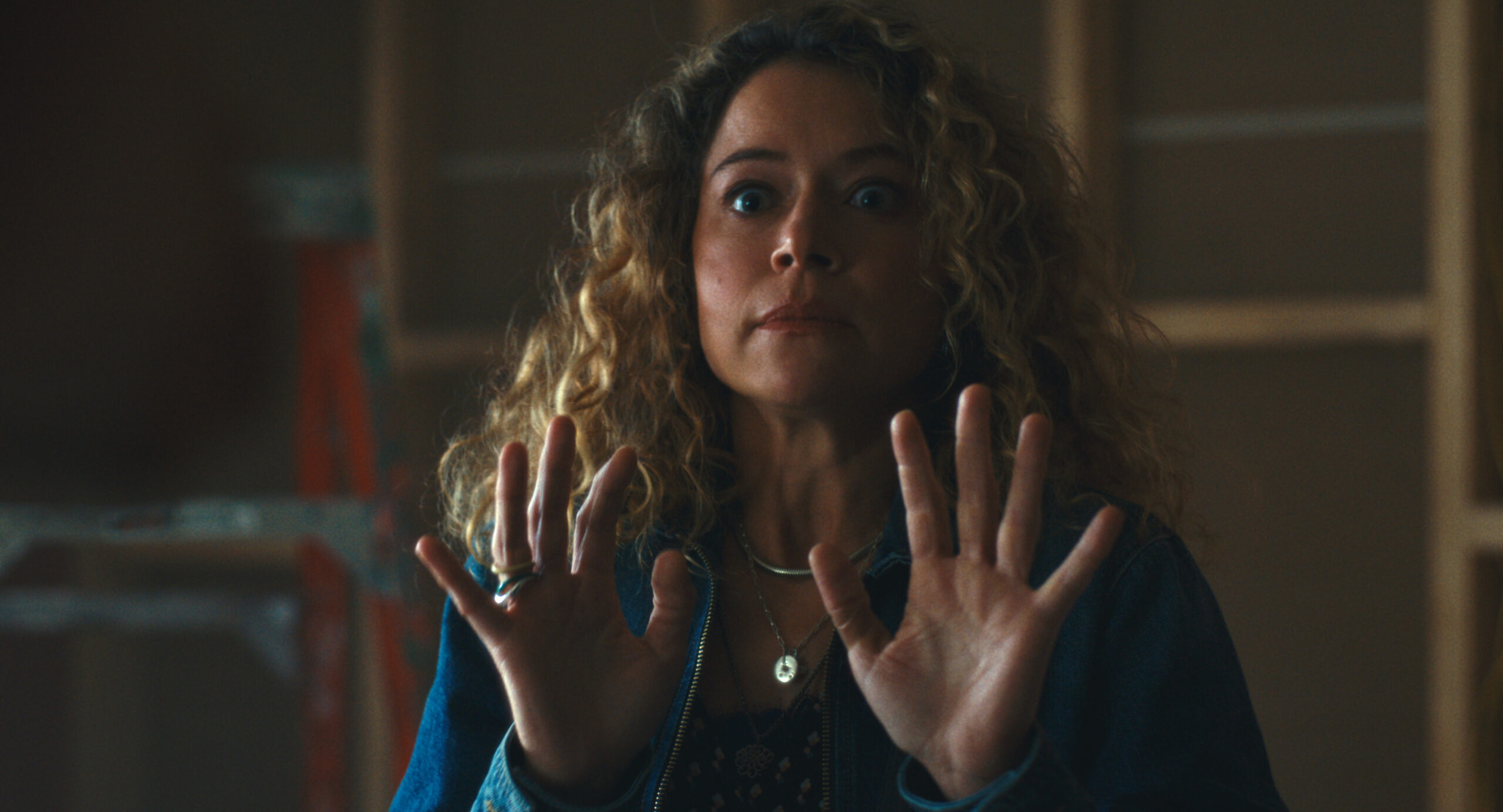 Maximum Pleasure Guaranteed Season 1 Episode 5 Review: Scamboy Woman with curly blonde hair raises both hands, eyes wide, in a dimly lit room, showing surprise or fear.
