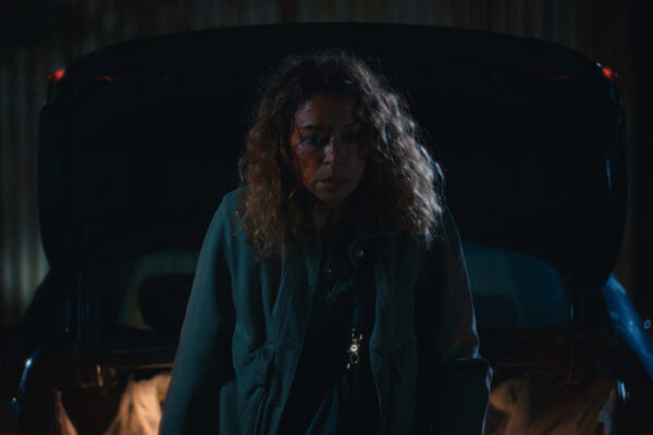 Woman with curly hair stands in front of a dark car in a dimly lit garage at night.