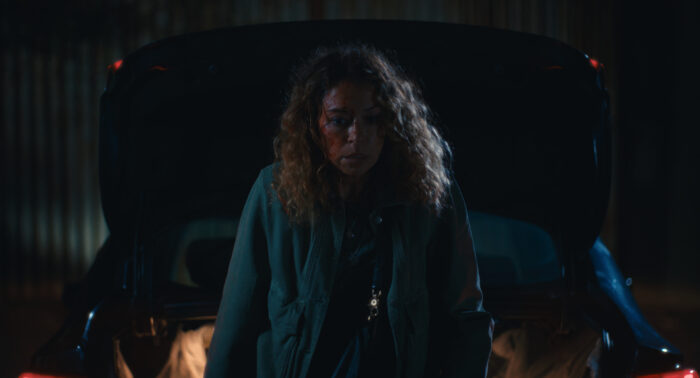 Maximum Pleasure Guaranteed Season 1 Episode 7 Review: Flighting Woman with curly hair stands in front of a dark car in a dimly lit garage at night.