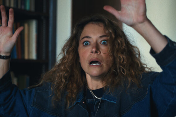 Maximum Pleasure Guaranteed Season 1 Episode 8 Review: Hallidays Woman with curly hair and a denim jacket raises her hands in surprise indoors, with bookshelves in the background.