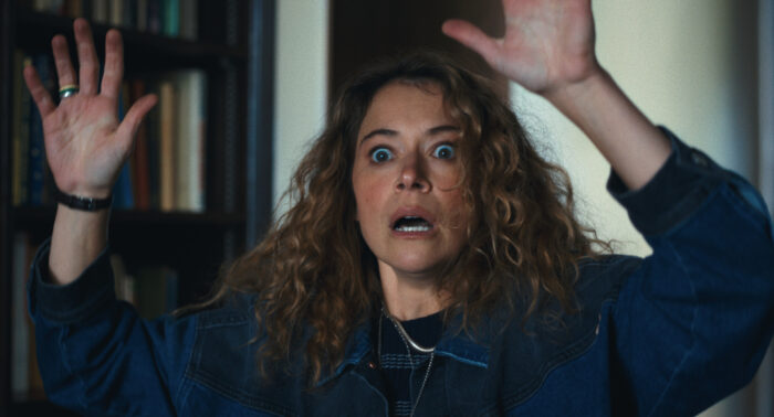 Maximum Pleasure Guaranteed Season 1 Episode 8 Review: Hallidays Woman with curly hair and a denim jacket raises her hands in surprise indoors, with bookshelves in the background.