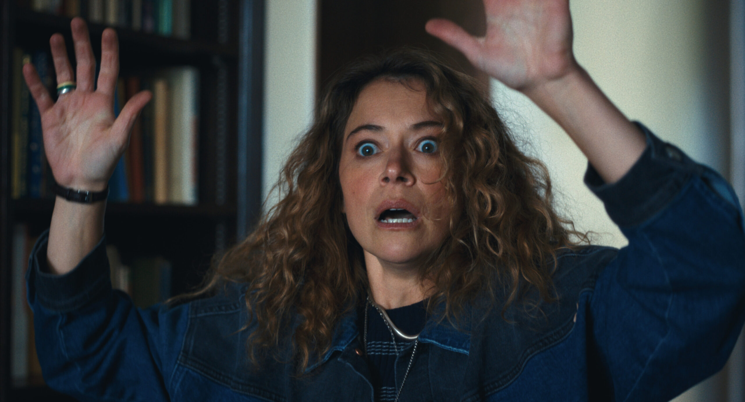 Maximum Pleasure Guaranteed Season 1 Episode 8 Review: Hallidays Woman with curly hair and a denim jacket raises her hands in surprise indoors, with bookshelves in the background.