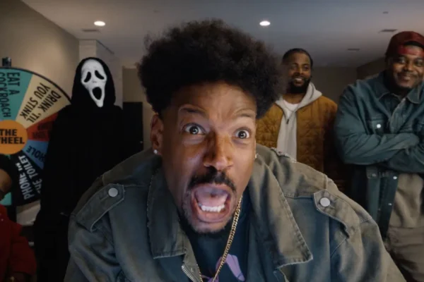 Close-up of a man in a denim jacket making a surprised, open-mouthed expression toward the camera, with friends and a ghost-face figure in the background and a colorful wheel behind them. - Scary Movie 6