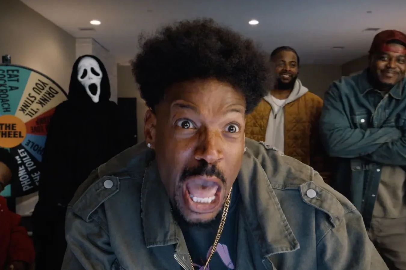 Scary Movie 6 Review: Only a Few Zany Scenes Hit the Comic Bullseye in This Wild Sequel Close-up of a man in a denim jacket making a surprised, open-mouthed expression toward the camera, with friends and a ghost-face figure in the background and a colorful wheel behind them. - Scary Movie 6