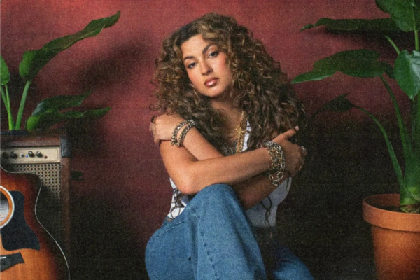 Tori Kelly Finds Strength, Faith, and Joy on God Must Really Love Me — Album Review Woman with curly hair sits cross‑armed in blue jeans, wearing chunky bracelets beside a guitar and amp in a retro room with plants.