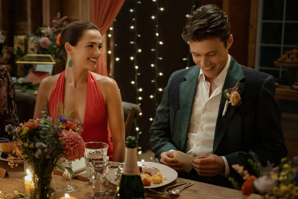 Voicemails for Isabelle Review: A Sweet Rom-Com That is Among the Very Best Films of the Year Couple at a festively lit dinner table: woman in a red halter dress smiling at a man in a dark blazer, flowers and candles around them.