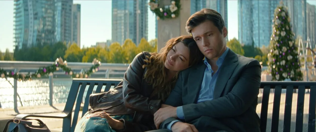 Voicemails for Isabelle Review: A Sweet Rom-Com That is Among the Very Best Films of the Year A couple sits close on a park bench by the water, leaning on one another with somber expressions in an urban waterfront setting.