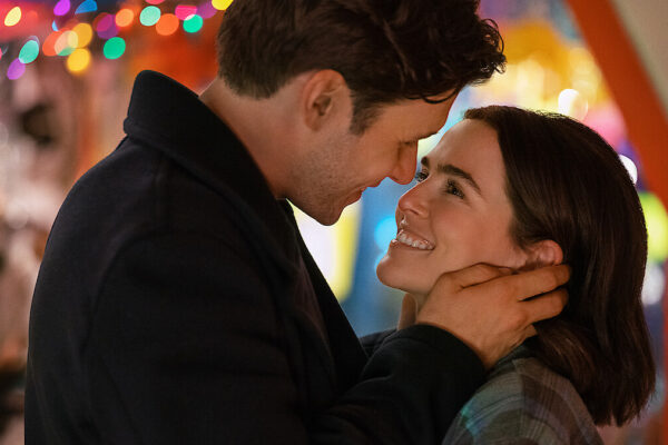A man tenderly holds a woman's face as they smile at each other under colorful holiday lights in the background.