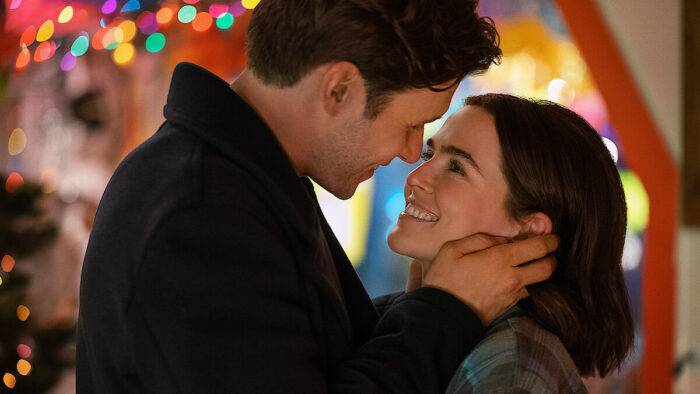 Voicemails for Isabelle Review: A Sweet Rom-Com That is Among the Very Best Films of the Year A man tenderly holds a woman's face as they smile at each other under colorful holiday lights in the background.