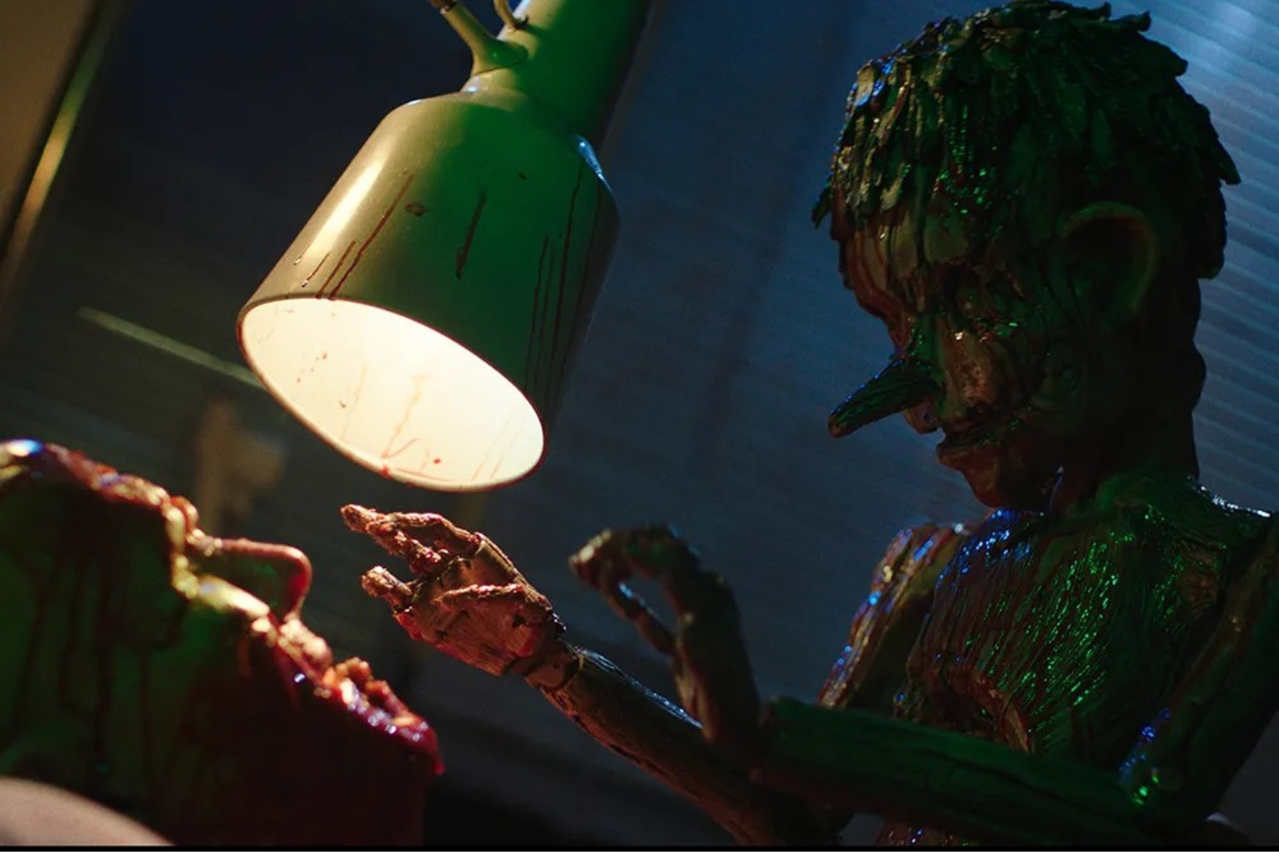 Pinocchio: Unstrung Preview: Release Date, Cast, Plot, and More Glossy green monster-like sculpture under a hanging lamp, with clawed hands reaching forward in a dimly lit scene.