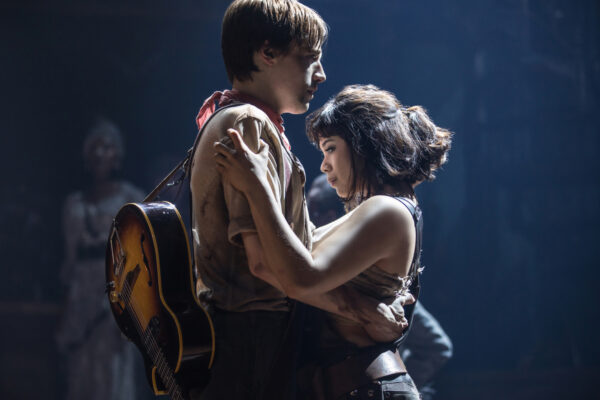 Two performers share an intimate embrace on stage, the man with a guitar slung over his back and a dusty shirt while the woman holds him close, bathed in moody stage light.