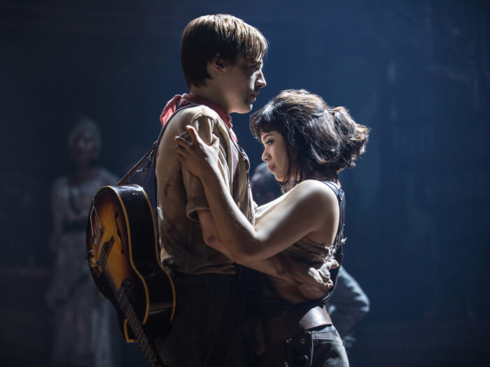 Hadestown: The Musical Preview: Release Date, Cast, Plot, and More Two performers share an intimate embrace on stage, the man with a guitar slung over his back and a dusty shirt while the woman holds him close, bathed in moody stage light.