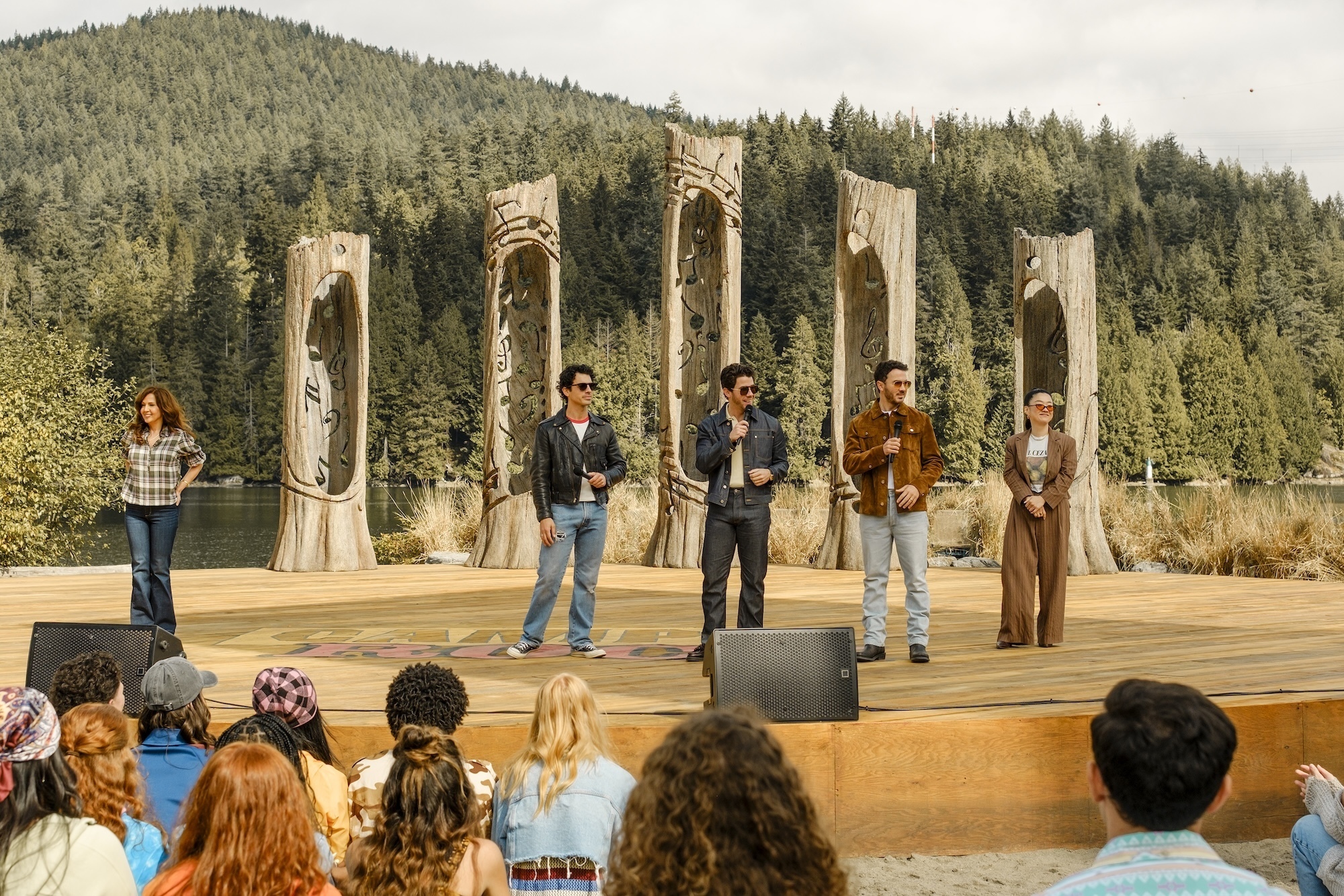 Camp Rock 3 Preview: Release Date, Cast, Plot, and More Five performers stand on an outdoor wooden stage, addressing a crowd, with tall carved posts and a forest backdrop behind them.