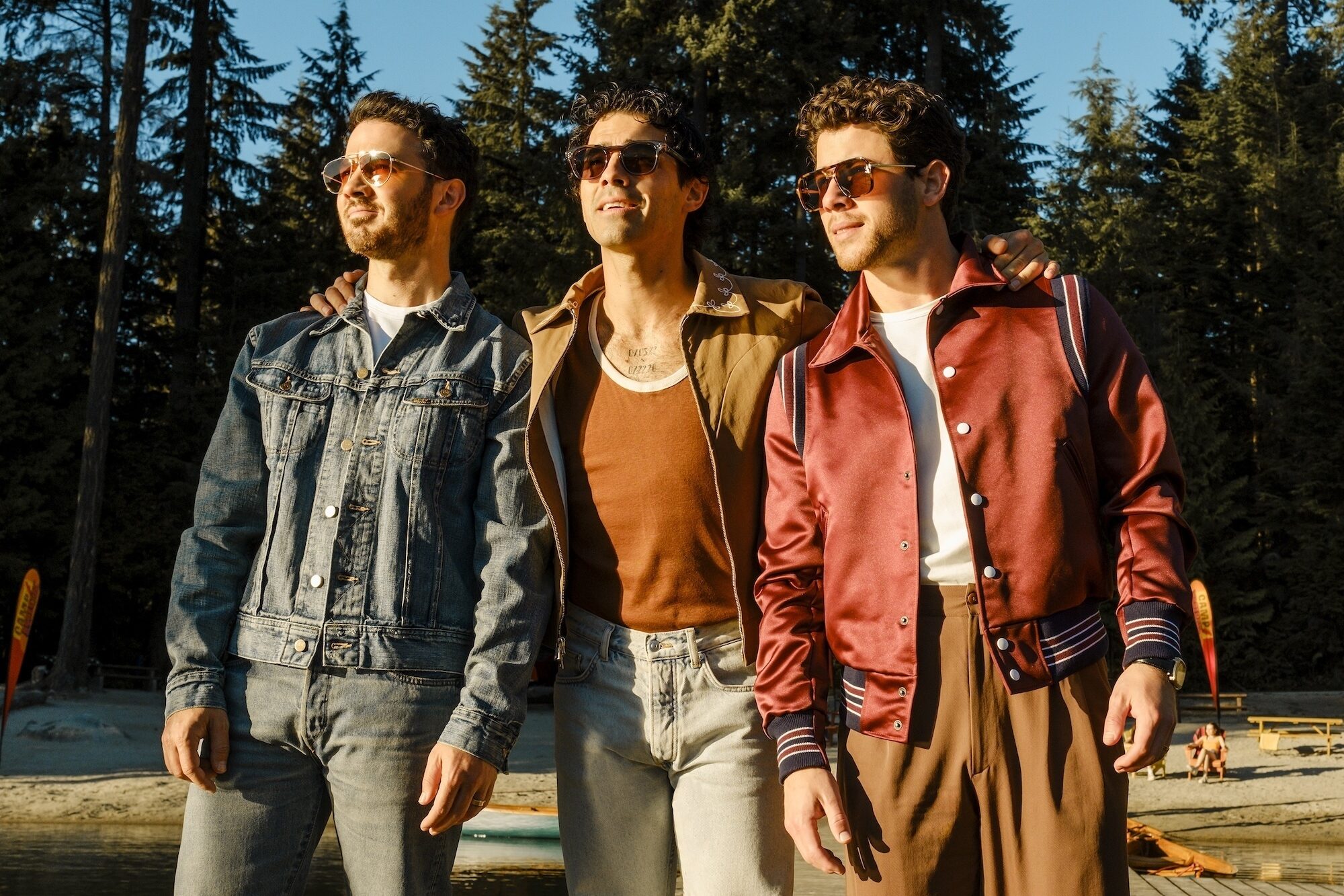 Camp Rock 3 Preview: Release Date, Cast, Plot, and More Three men wearing sunglasses pose together outdoors by a lake with tall trees in the background.