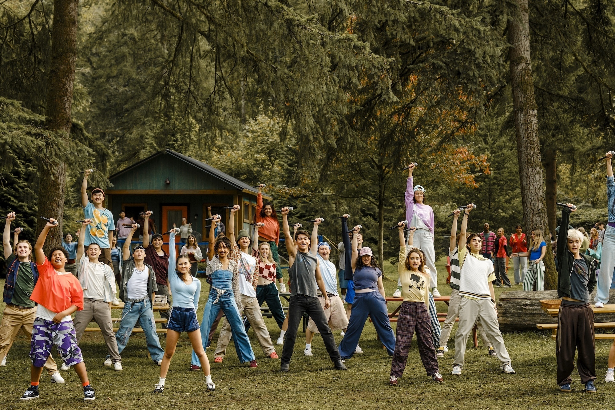 Camp Rock 3 Preview: Release Date, Cast, Plot, and More Large diverse group outdoors raising their arms in a synchronized activity, with trees and a cabin in the background (campground scene).