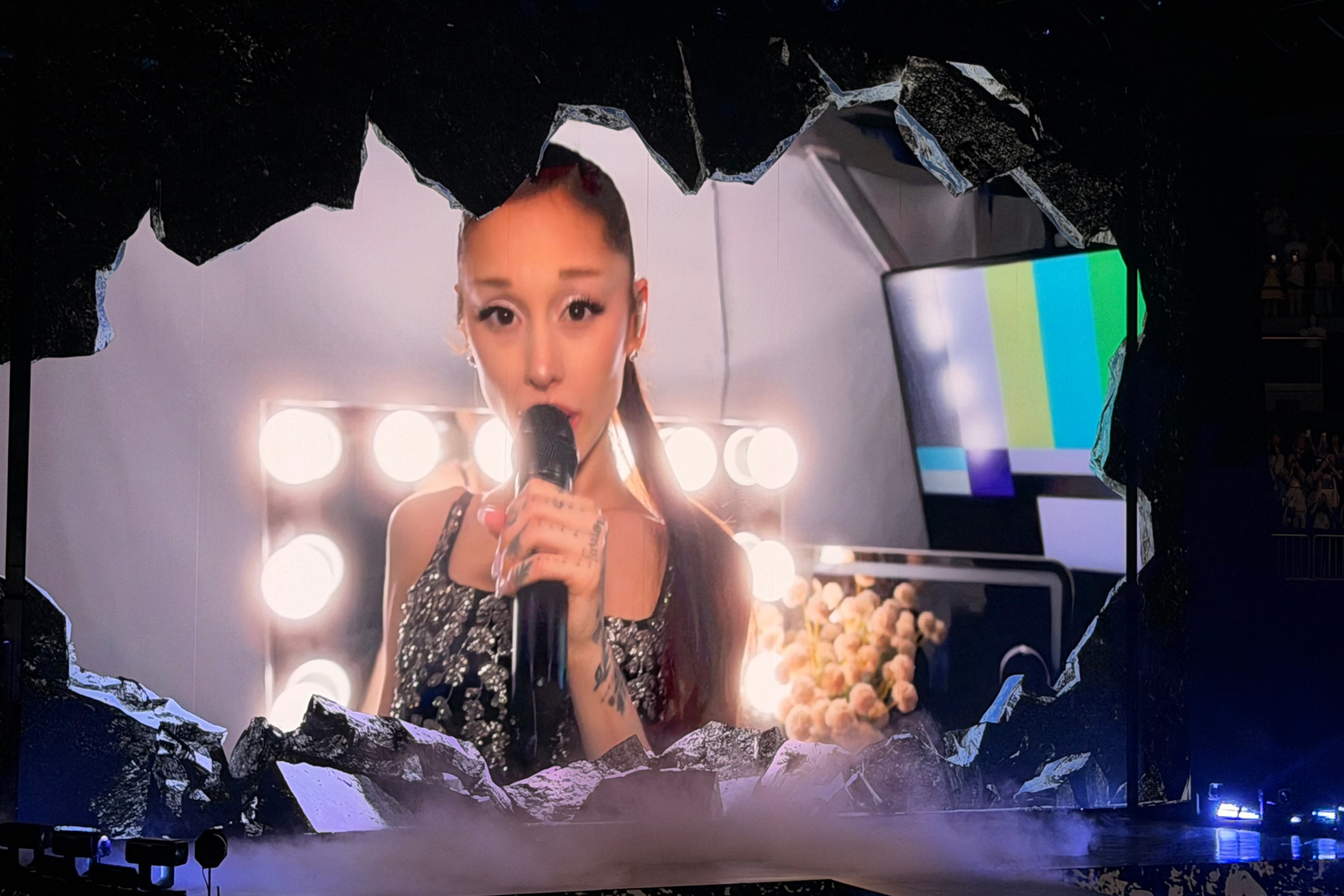 Ariana Grande’s Eternal Sunshine Tour Is Pure Magic Singer performs on stage, holding a microphone, with bright backstage lights and a torn-frame backdrop around her.