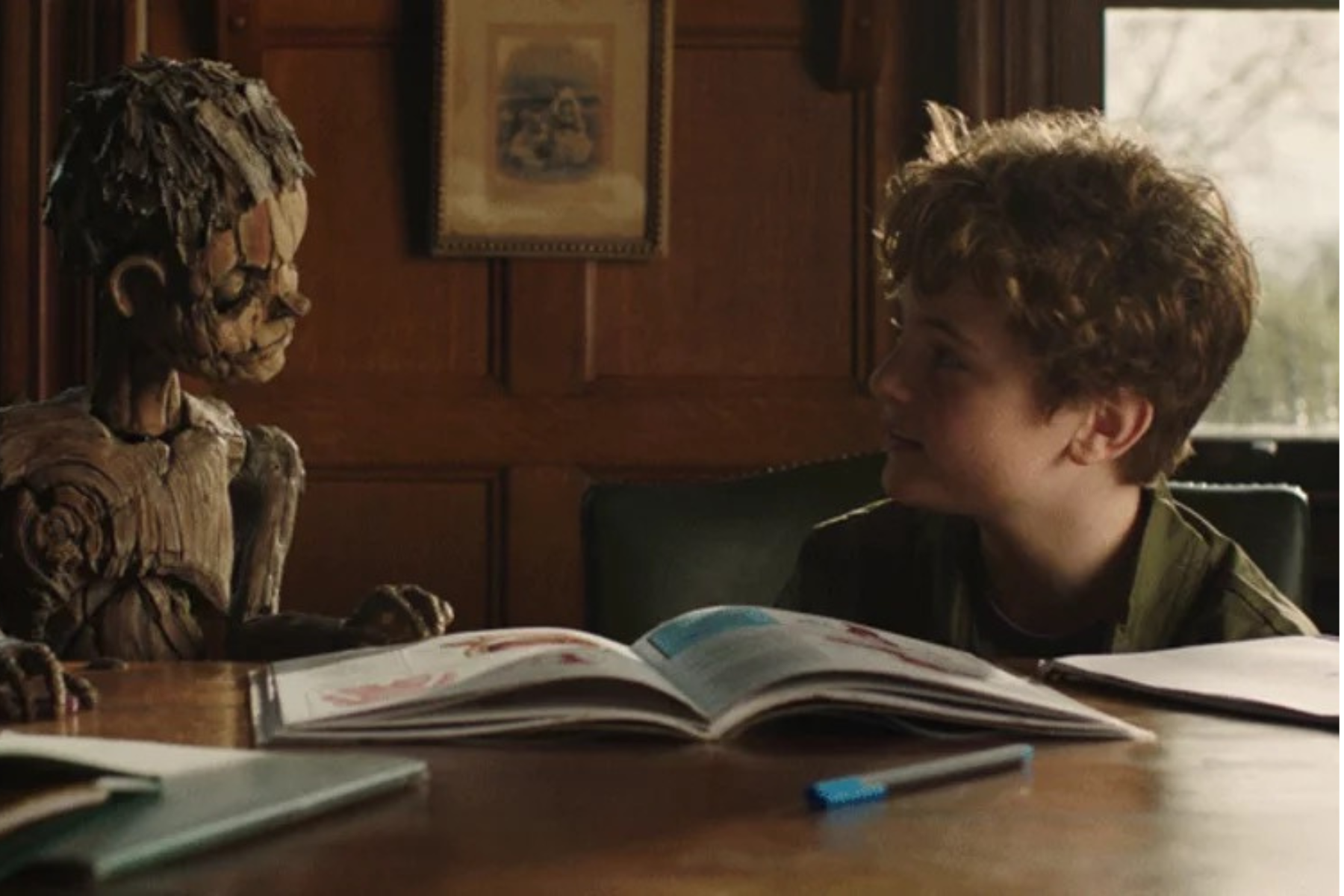 Pinocchio: Unstrung Preview: Release Date, Cast, Plot, and More Boy sitting at a wooden table, reading an open illustrated book beside a carved wooden statue in a warmly lit room.