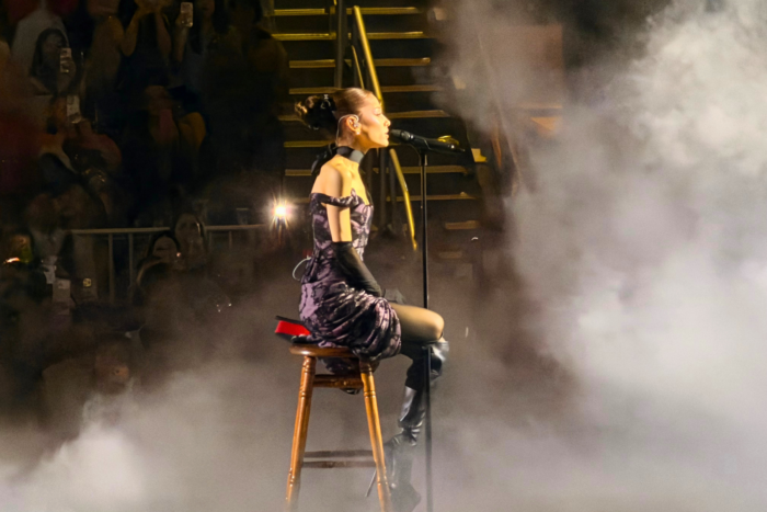 Ariana Grande’s Eternal Sunshine Tour Is Pure Magic Singer in a purple patterned dress sits on a wooden stool and sings into a microphone amid stage fog.