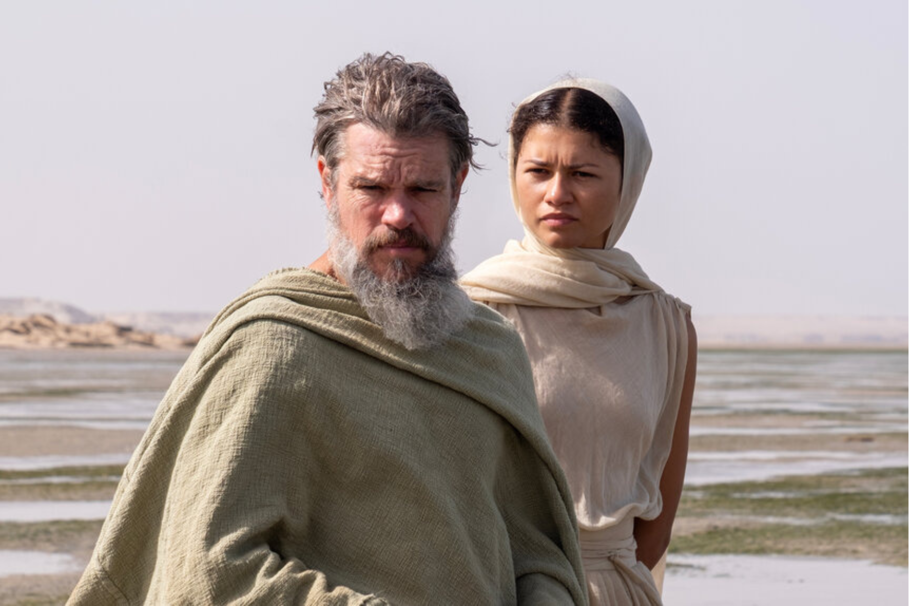 The Odyssey Review: Christopher Nolan Delivers a Modern Masterpiece Two people in light robes stand on a shoreline, with a bearded man in the foreground and a woman in a headscarf behind him, gazing ahead.