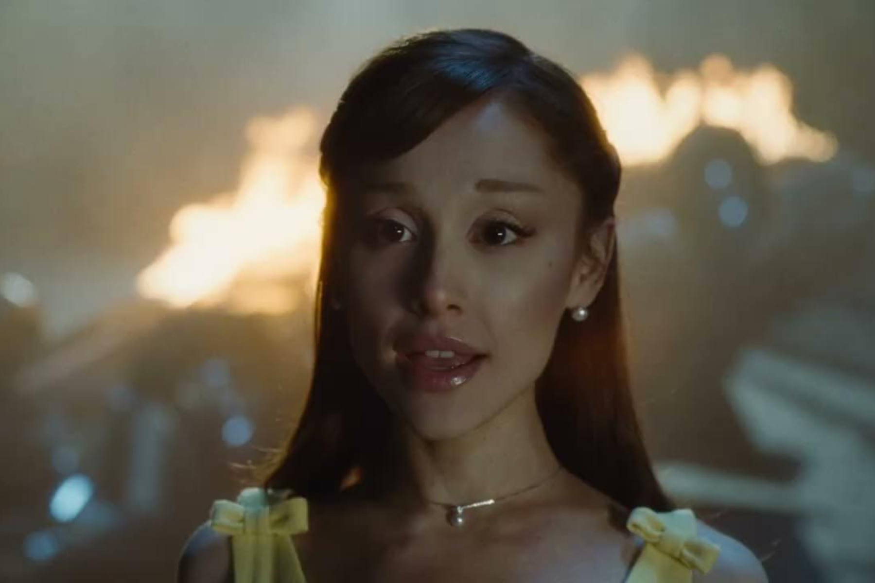 Ariana Grande Finds Power in Reinvention on petal — Album Review Close-up of a smiling woman wearing a yellow dress with bow straps, glowing city lights behind her.