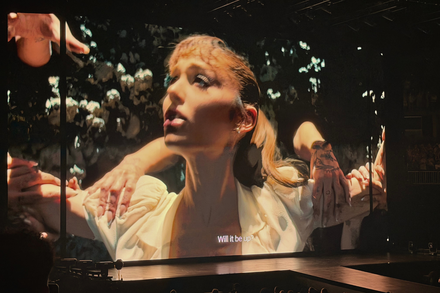 Ariana Grande’s Eternal Sunshine Tour Is Pure Magic Large projection of a woman on stage, looking up, with hands reaching toward her and a subtitle reading'Will it be up?'.