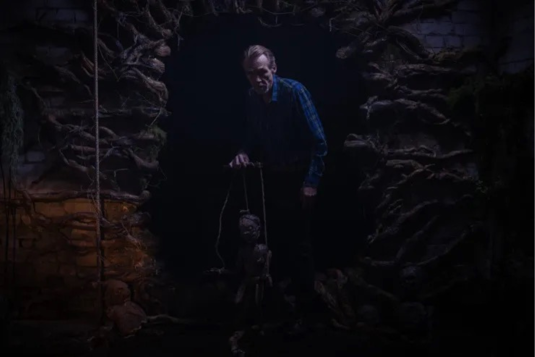 Pinocchio: Unstrung Preview: Release Date, Cast, Plot, and More A man in a blue plaid shirt stands in a dark stone dungeon, holding a rope attached to a hanging prop head.