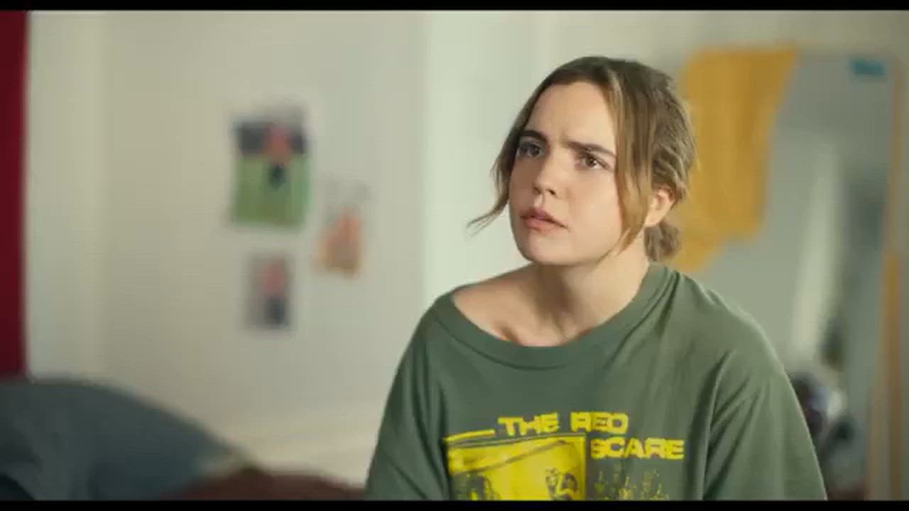Joel Courtney and Andy Delaney Talk 40 Dates and 40 Nights, Dating Red Flags, and Empathy Woman wearing an olive-green T-shirt with a yellow graphic that says 'THE RED SCARE,' looking puzzled in a casual indoor setting.