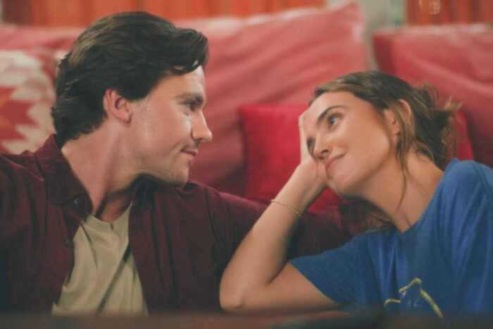 Joel Courtney and Andy Delaney Talk 40 Dates and 40 Nights, Dating Red Flags, and Empathy A couple lying on a red couch facing each other, smiling softly and sharing a warm moment.