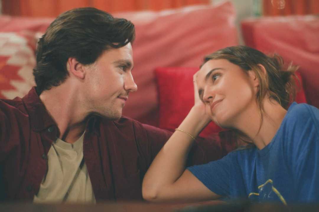 Joel Courtney and Andy Delaney Talk 40 Dates and 40 Nights, Dating Red Flags, and Empathy A couple lying on a red couch facing each other, smiling softly and sharing a warm moment.