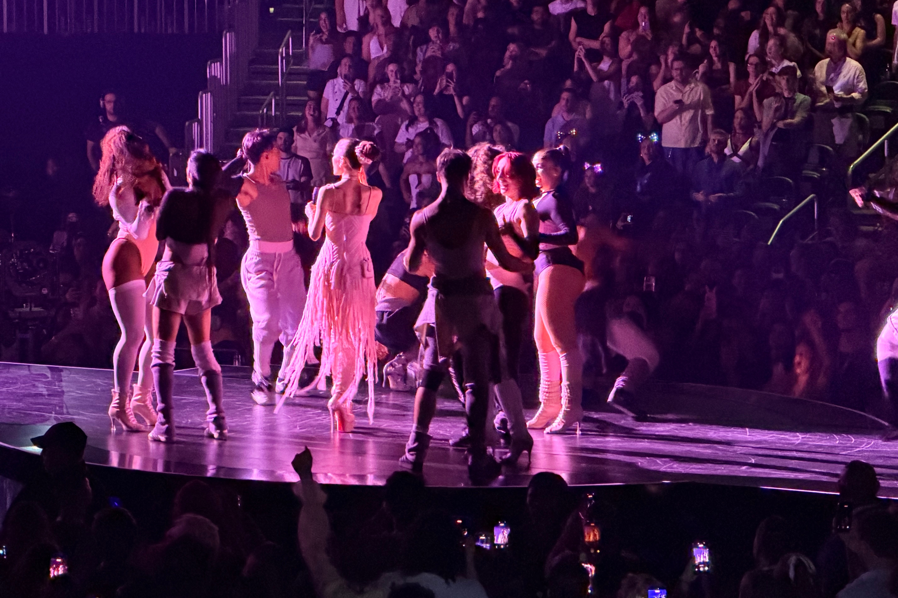 Ariana Grande’s Eternal Sunshine Tour Is Pure Magic Group of performers in white outfits on a lit stage, dancing while a cheering audience watches in a dimly lit arena.