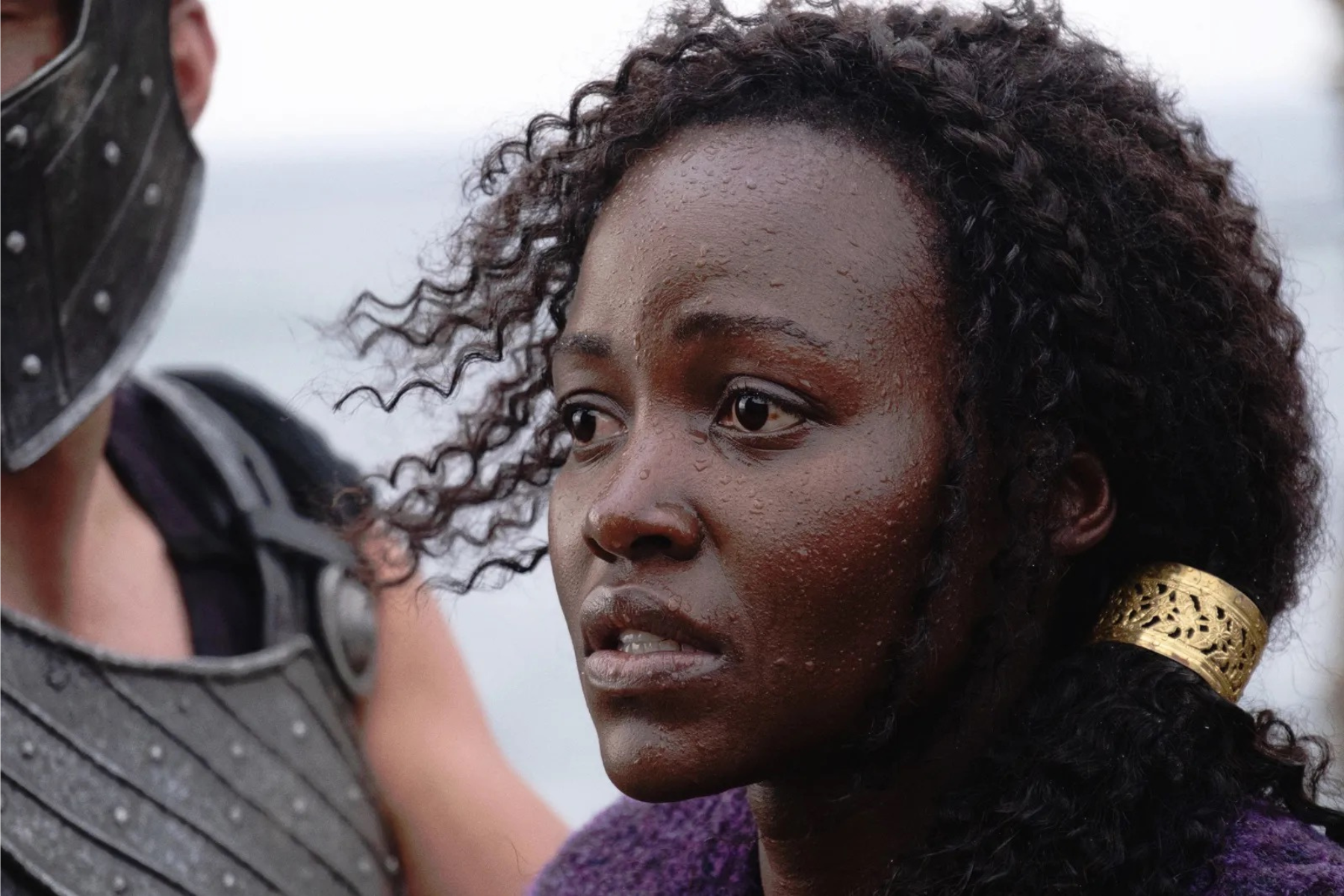 The Odyssey Review: Christopher Nolan Delivers a Modern Masterpiece Close-up of a young Black woman with curly hair, a gold earring, and droplets on her skin, beside a person wearing armor.