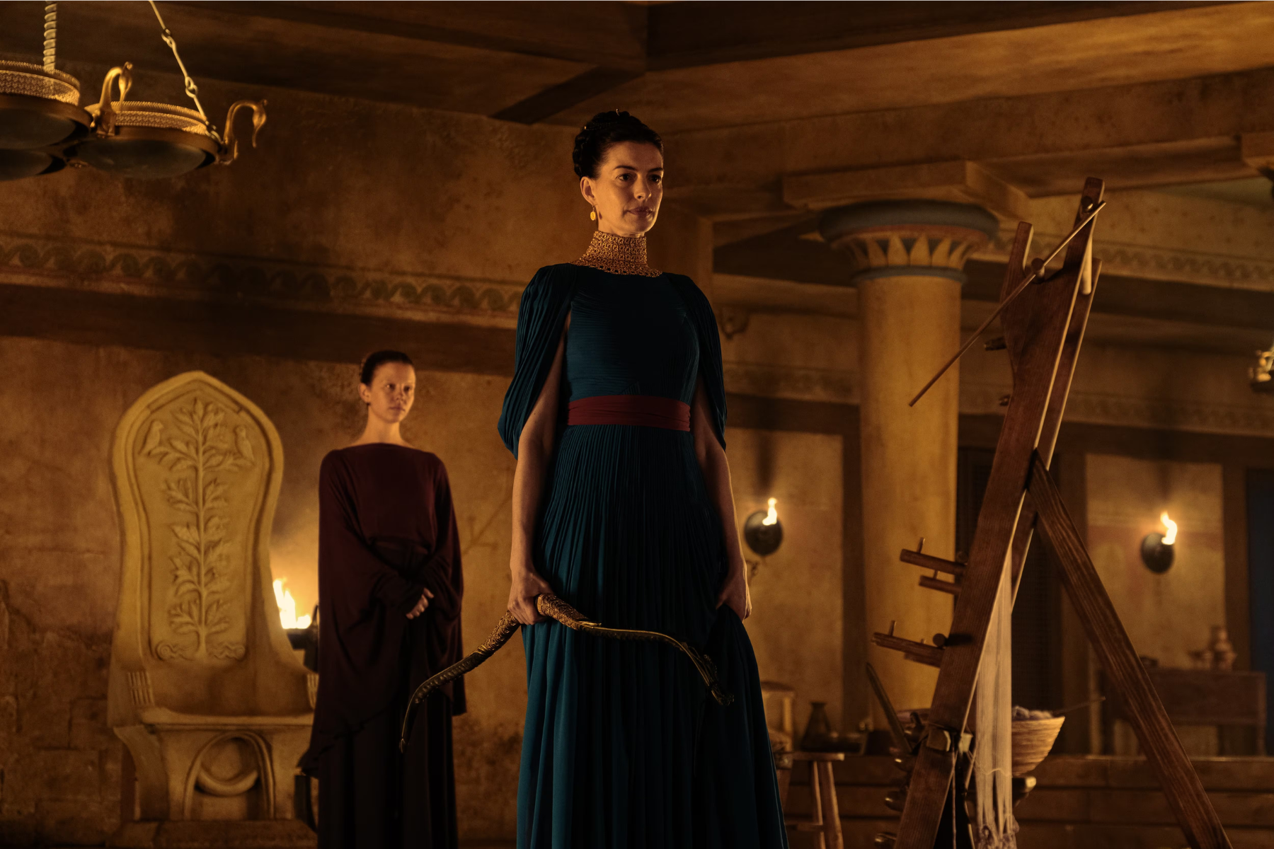 The Odyssey Review: Christopher Nolan Delivers a Modern Masterpiece Central woman in teal gown with gold choker holding a curved sword in a dim torchlit, ancient-style room with another woman in burgundy in the background.