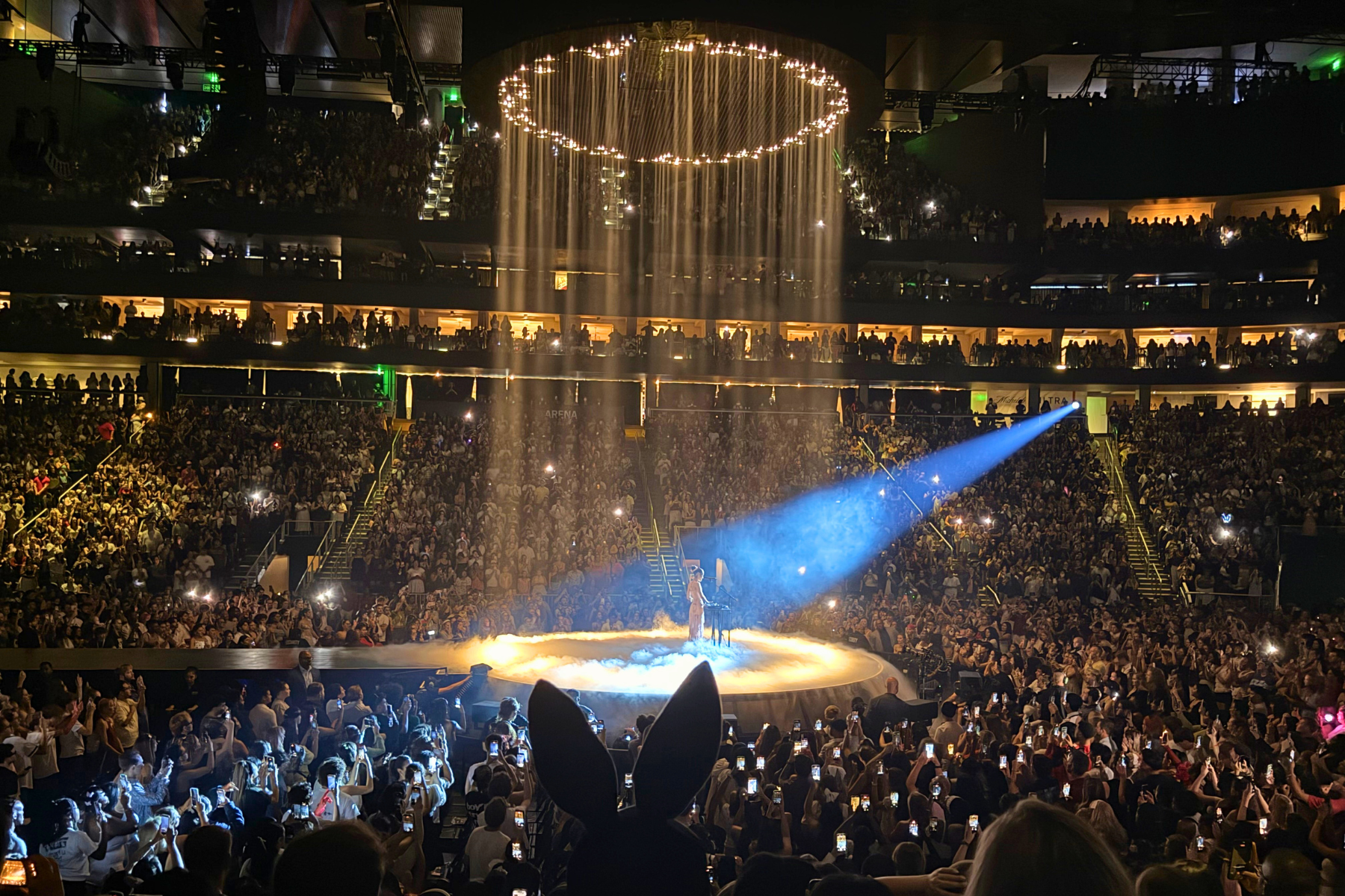 Ariana Grande’s Eternal Sunshine Tour Is Pure Magic Crowd-filled multi-tier arena watching a performer on a circular, halo-lit stage with warm overhead lights and fog.