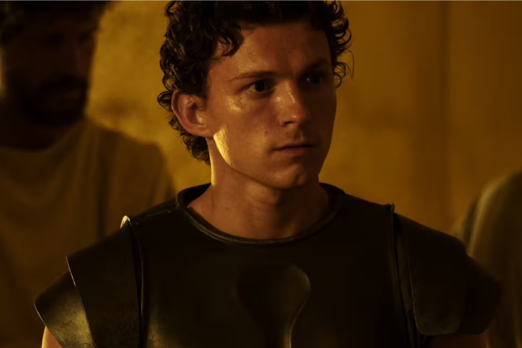 The Odyssey Review: Christopher Nolan Delivers a Modern Masterpiece Close-up of a young man in bronze armor, looking off to the right in a dim amber-lit scene.